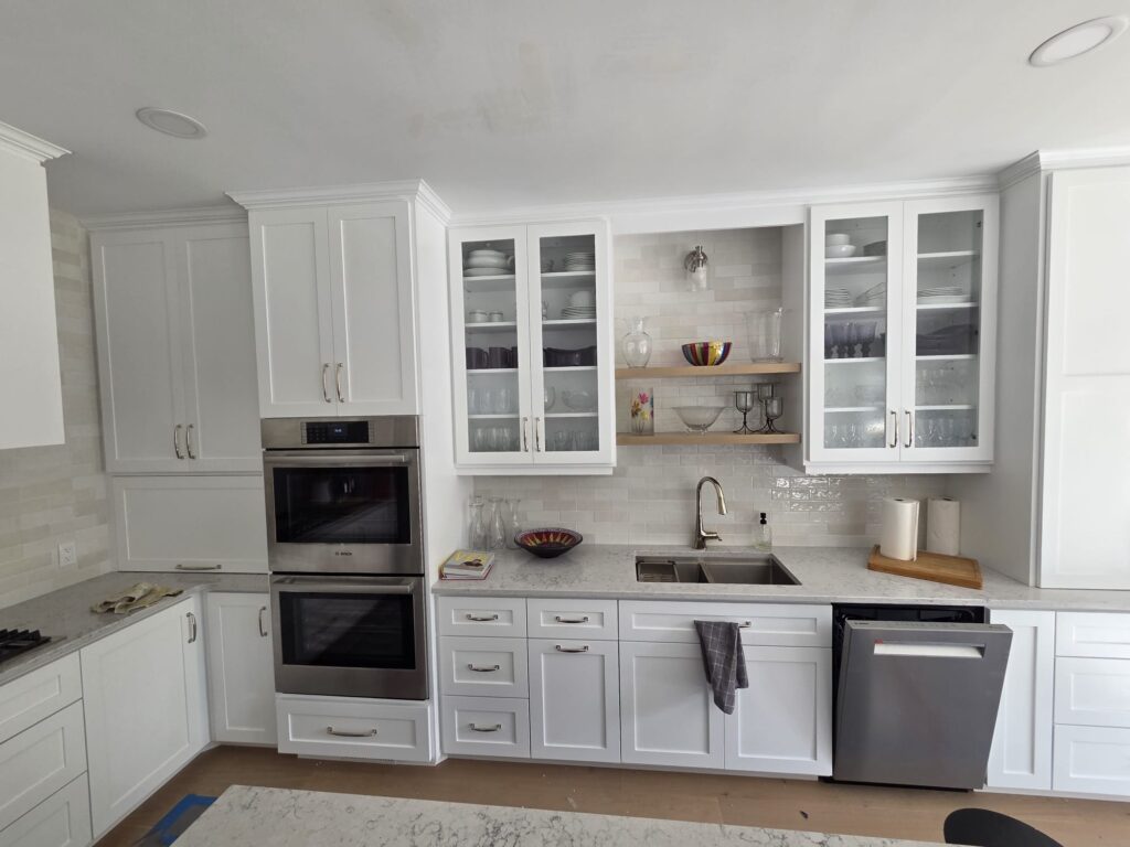 white kitchen with double oven and a sink in the center with glass door upper cabinets and floating shelves.